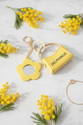 LE MIMOSA – Bijou de sac porte-clé jaune sur marbre blanc entouré de fleurs de mimosa, fleur miroir et mini sac cuir, DELPHINUS