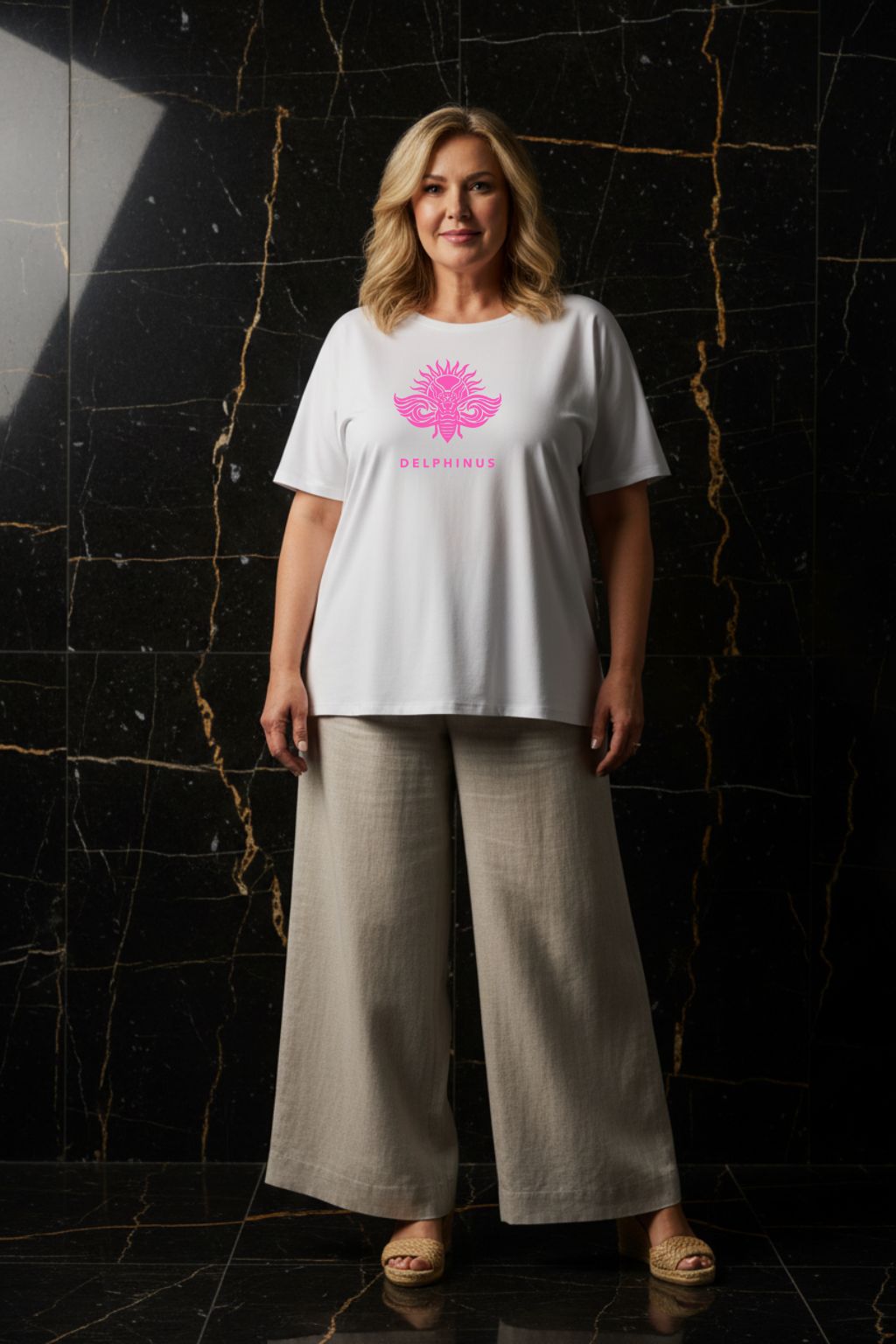 LA PÉTILLANTE CURVE – T-shirt unisexe blanc logo rose DELPHINUS, femme curve mature et élégante, ambiance premium marbre noir et or, pantalon large et espadrilles, DELPHINUS