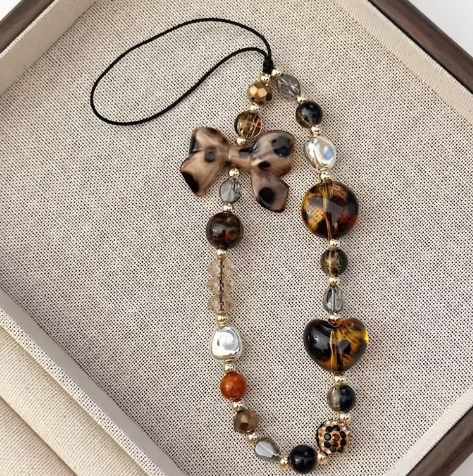 Bijou de téléphone – perles brunes, nacrées et dorées avec nœud et cœur effet écaille, DELPHINUS