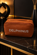 L'ICONIQUE – trousse de toilette unisexe en similicuir marron cognac vue de face sur socle noir aux finitions dorées, DELPHINUS