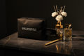 Pochette posée sur marbre, vue de face, accessoires dorés L'Intrépide DELPHINUS — pochette oxford noire inscription DELPHINUS en blanc, posée sur marbre noir, entourée d'un diffuseur fleurs blanches, d'un flacon de parfum et d'un stylo dorés. Élégance et praticité réunies.